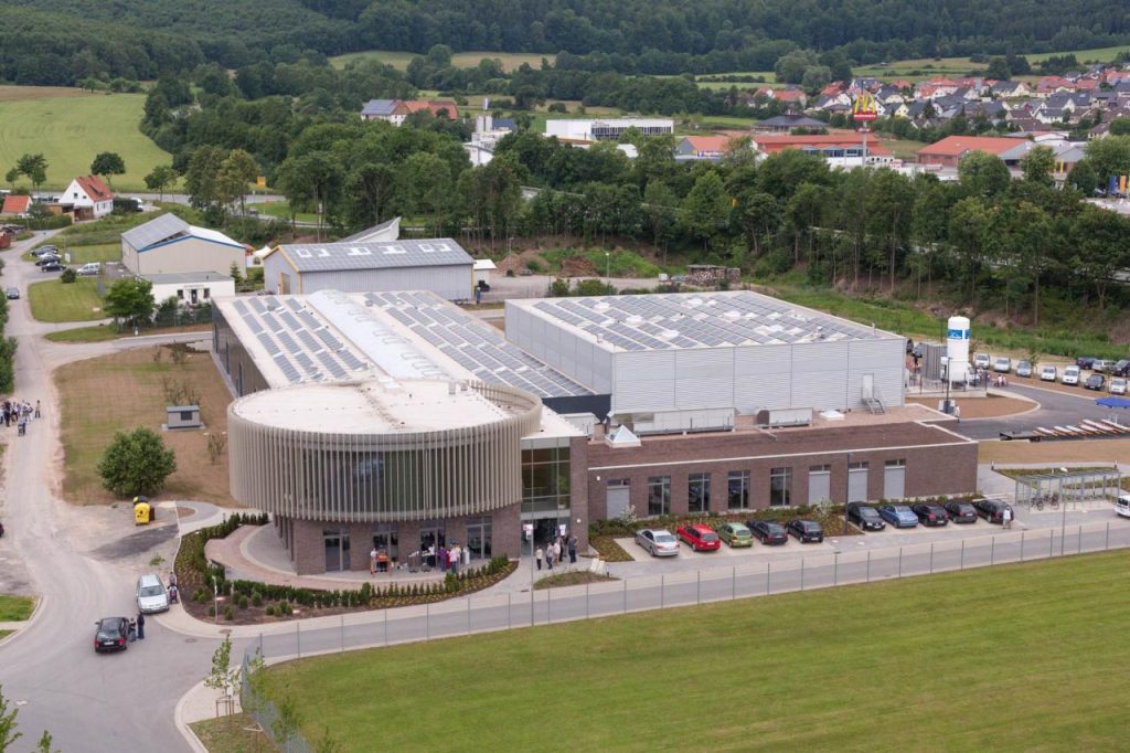 Luftaufnahme des Firmengebäudes Integra EG mit Produktionshalle und Bürotrakt. An der Ecke ein prägnanter runder Bereich mit Holz verkleidet davor parkende Autos