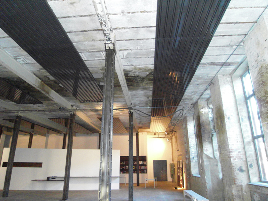 Industriell gestalteter Innenraum mit sichtbarer Betondecke, schwarzen Deckenheizflächen und Stahlstützen