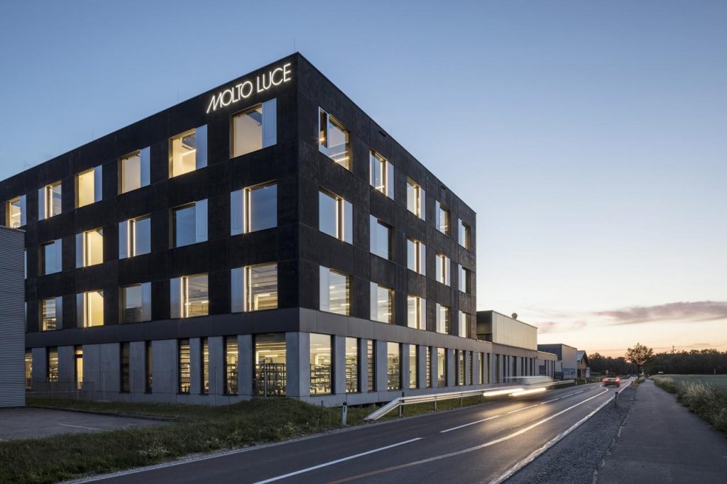 Außenansicht des Molto Luce Firmengebäudes mit beleuchteten Fensterfronten bei Abenddämmerung