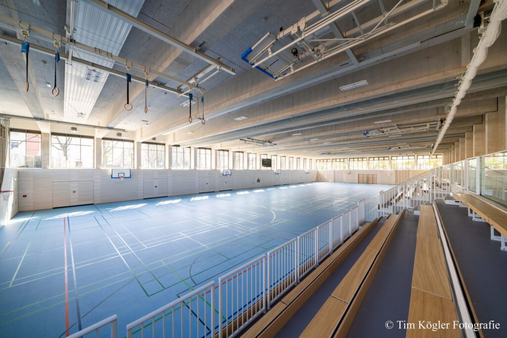 Moderne Dreifach-Sporthalle mit hellblauem Boden, bunten Spielfeldmarkierungen und fest installierten Trib&uuml;nen, gro&szlig;e Glasfronten lassen viel Tageslicht herein.